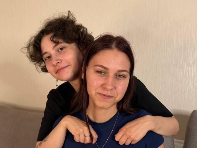 TaraCastongvay, avec ses cheveux bruns bouclés et sa petite poitrine, se trouve derrière TaraCastongvay, qui a les cheveux roux lisses et un joli visage. Toutes les deux portent des t-shirts noirs et bleus et sourient..