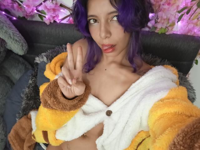 DarckVelvet with purple hair, yellow and brown fuzzy jacket, white shirt, showing a peace sign, small tits, light skin, pink lips, sitting on a gray couch.