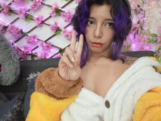 DarckVelvet with purple hair, showing a peace sign, wearing a yellow and brown fuzzy jacket with a white button-up, exposing her tits. Pink fairy lights in the background.