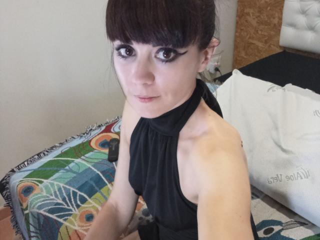 OrianaLaFrancaise, com cabelo castanho escuro, vestido preto de alças e maquiagem marcante, sentada em uma cama colorida. Seus pequenos seios são visíveis. Ela está olhando para a câmera..