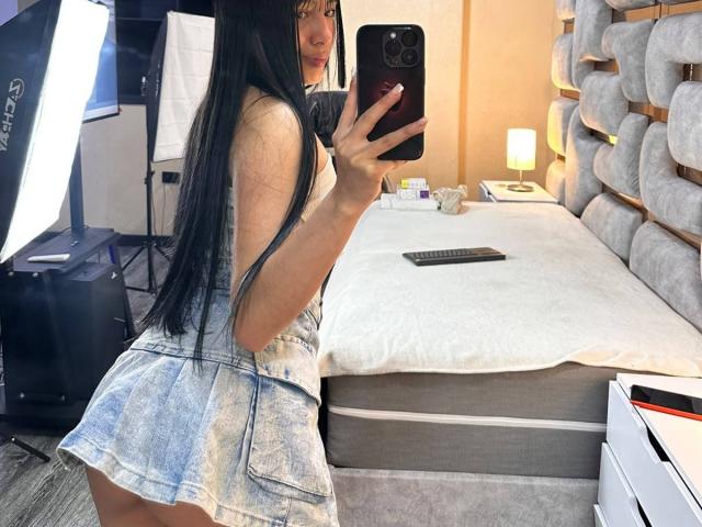 SkarletAry, con lunghi capelli neri, indossa un vestito di jeans e sta scattando un selfie allo specchio, mostrando il suo sedere. Tiene in mano un telefono nero, in una camera da letto con un letto..