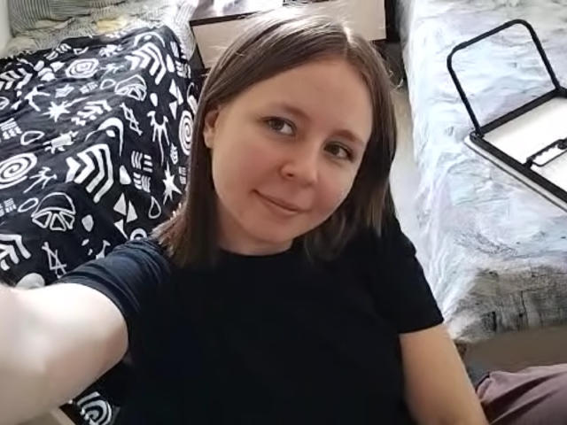 Johnetta Urtiaga, met lichte huid, bruin haar, een zwart shirt, lachend naar de camera, kleine borsten, een bed met een zwart-wit geborduurde deken, een wit laken, een zwarte make-up tray op de achtergrond..