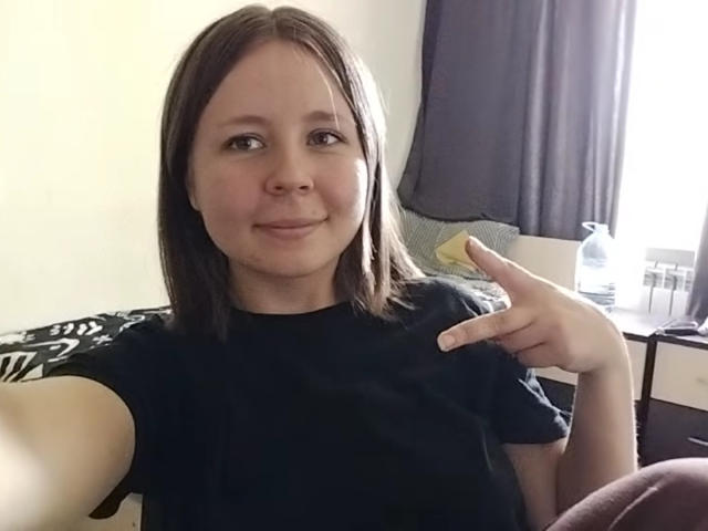 Johnetta Urtiaga, met halflang bruin haar, een zwart T-shirt en lichte huid, glimlacht licht en wijst met haar rechter wijsvinger, in een informele, binnenomgeving..