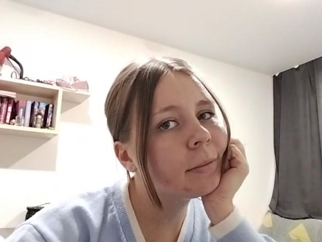 Johnetta Urtiaga met bruin haar in een staart, een blauwe trui, een klein puistje aan de rechterwang, met haar kin op haar hand rustend, zacht glimlachend, witte muren, een plank met flessen, een grijze gordijn..