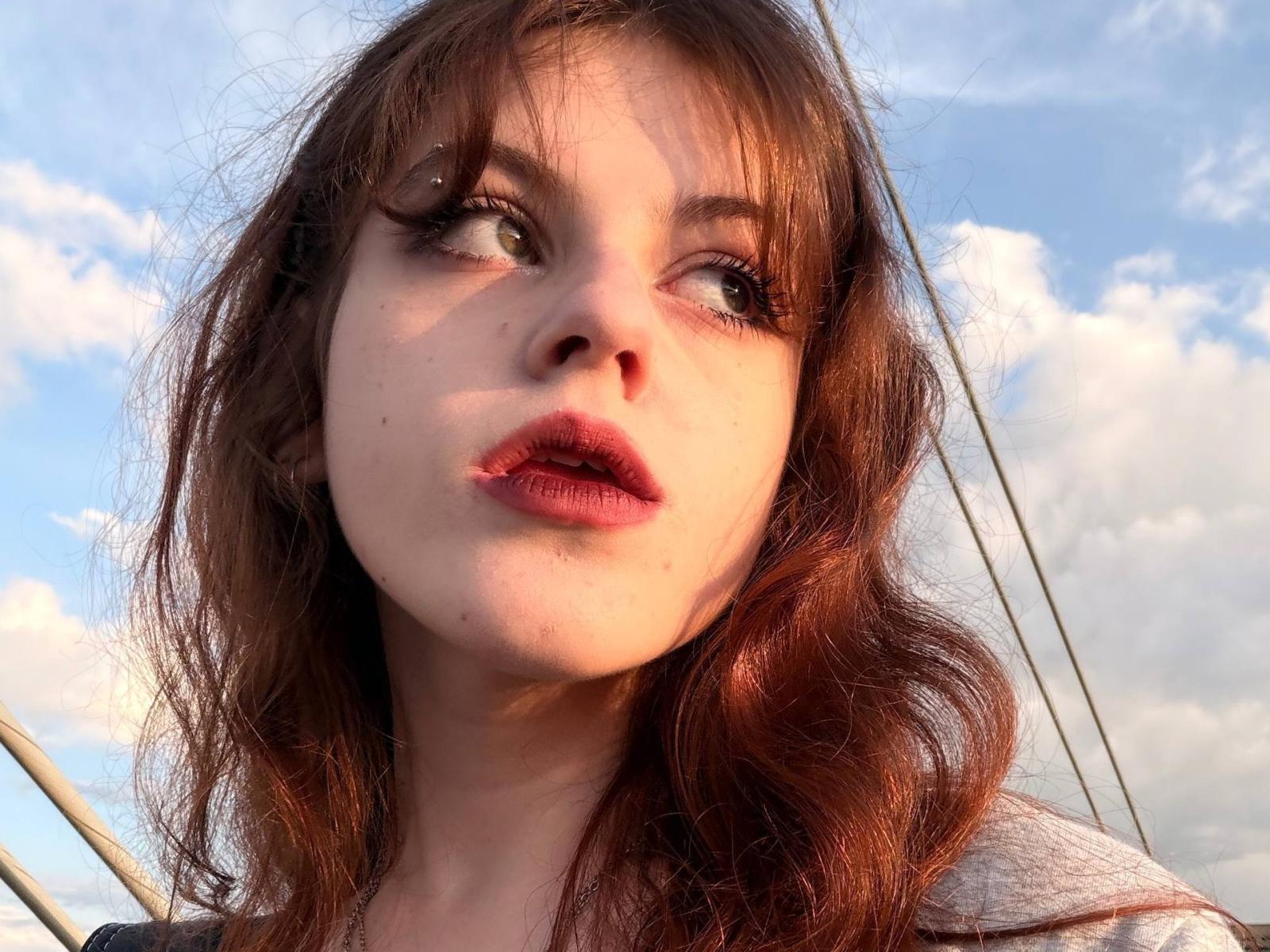 ArvillaUptmor tem cabelos ondulados, castanho-avermelhados, pele clara e lábios rosados com um leve brilho. Ela está olhando para cima com os olhos semicerrados, mostrando uma maquiagem discreta e delineador escuro..