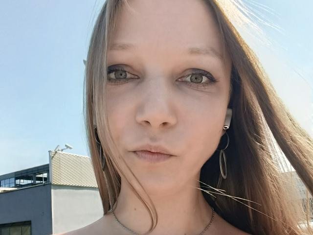 LovelyxEmilly hat hellbraunes Haar, grüne Augen und einen blassen Hautton. Sie trägt kleine Reifenohrringe und eine feine Halskette und hat einen neutralen Gesichtsausdruck..