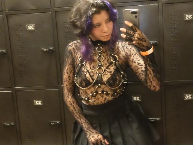 DarckVelvet with purple hair, wearing a lace top with metal chains over her tits, black skirt, and fingerless gloves, taking a mirror selfie in a locker room.