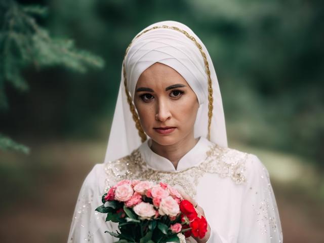 Donna con hijab bianco, che tiene un bouquet di rose rosa e rosse, indossa un abito bianco con ricami dorati, espressione seria, sfondo verde sfocato, luce soffusa, guarda direttamente verso la telecamera..
