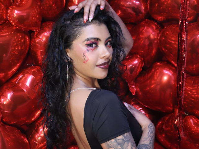 DevilAliceXo met krullend zwart haar, rode oogmake-up en tatoeages, gekleed in een zwarte top, staand voor rode, hartvormige ballonnen, met haar hand in haar haar, en om zich heen kijkend..
