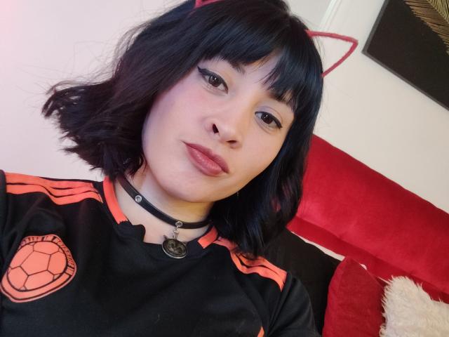 IssisDiosa, con cabello negro, orejas de gato, collar negro, delineador de ojos, labios rojos, camisa negra con detalles naranjas y un emblema de tortuga, sentada en un sofá rojo, mirando a la cámara..