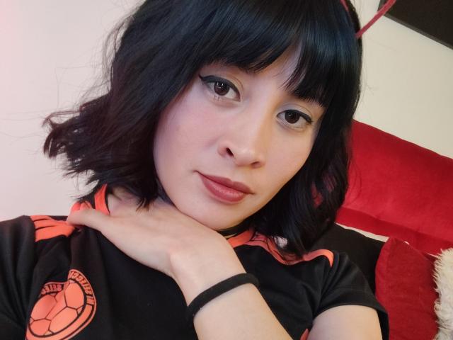 IssisDiosa, com cabelo preto ondulado, lábios vermelhos e camisa de futebol preta e laranja, apoiando o queixo na mão, olhando para a câmera com um leve sorriso..