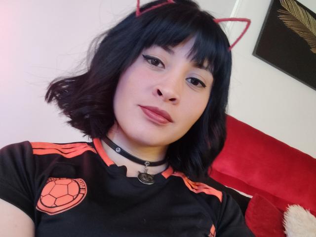 IssisDiosa com orelhas de gato pretas, cabelo preto ondulado, colar preto, camisa de futebol vermelha e preta, batom vermelho e um logotipo de bola de futebol na camisa, sentada em uma cadeira vermelha..