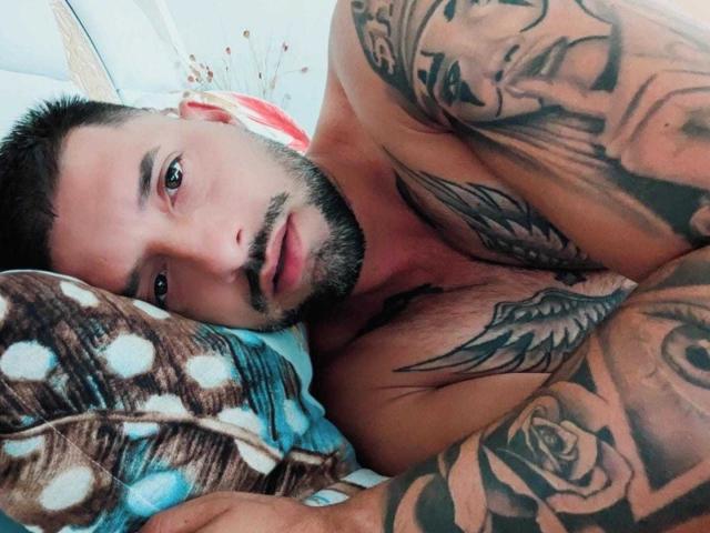 TommyDum, sin camisa y con tatuajes, está acostado sobre un cojín con diseños azules y marrones. Tiene barba, cabello oscuro y corto, y una expresión seria, mirando a la cámara..