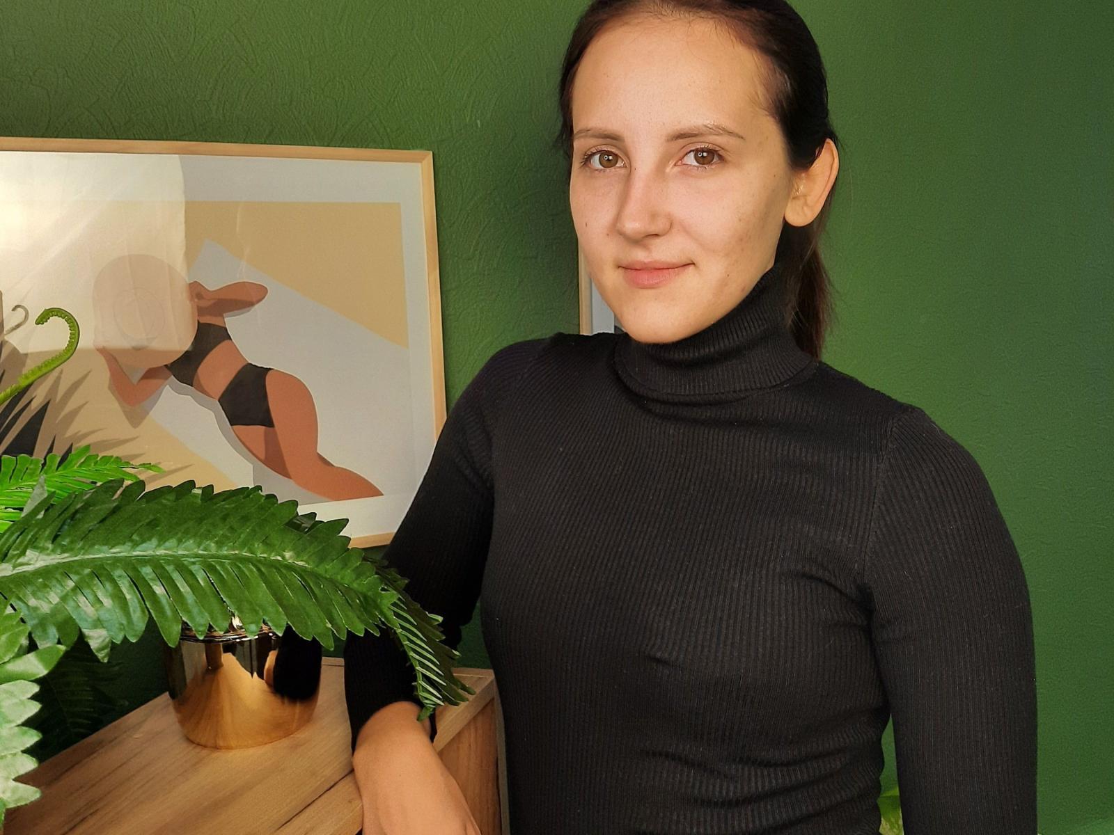 ElmaChochrek, avec des cheveux bruns et portant un pull noir à col roulé, se tient près d'une plante verte et d'un meuble en bois, souriant à l'appareil photo, avec un tableau abstrait encadré en arrière-plan..
