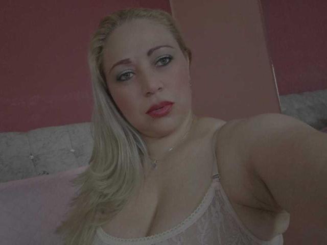 MariaRousell with blonde hair, blue eyes, red lipstick, wearing sheer white top, exposing her tits, arm raised, looking at camera, pink and gray background.