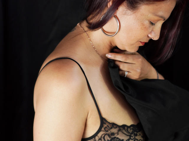 MilenneSexy tem a pele clara, cabelos ruivos e veste um sutiã de renda preto, mostrando seus seios. Ela está puxando uma camisa preta, revelando um anel no dedo..