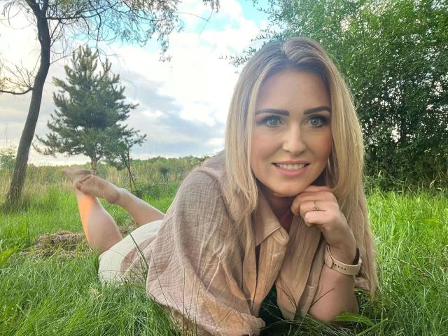 HotMarie-hot with blonde hair, lying in grass, wearing a pink shirt, smiling at camera, showing a cute face, her ass up, hands near her mouth, ring on finger.