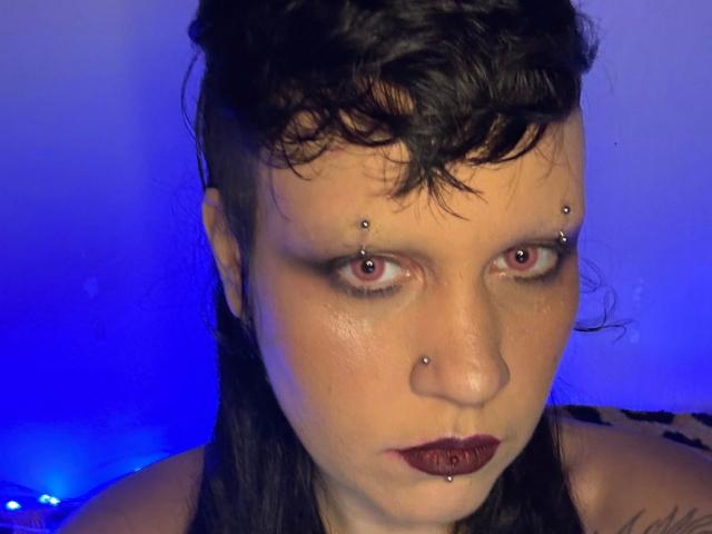 Kaotyka-hot has dark, wavy hair, piercings in both eyebrows and nose, red eyes, and dark red lipstick. Blue light highlights her pale skin. She's looking directly at the camera.