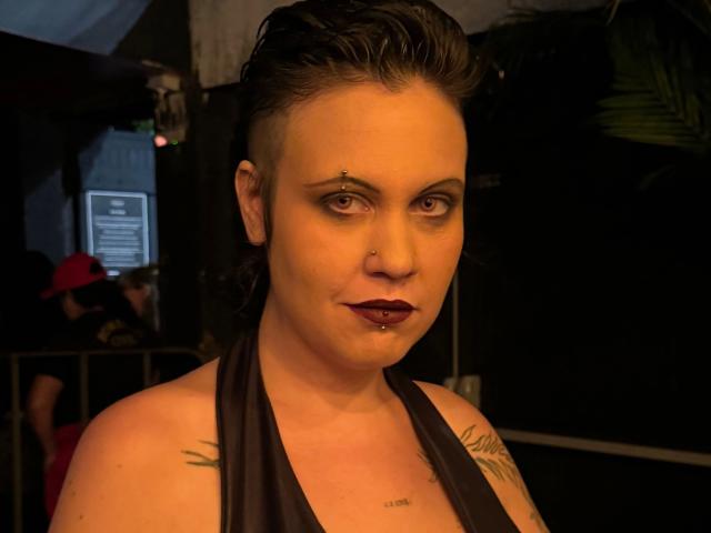 Kaotyka-hot has a shaved side, dark lipstick, nose ring, lip ring, and multiple tattoos. She's wearing a black halter top, looking directly at the camera with a slight smirk.
