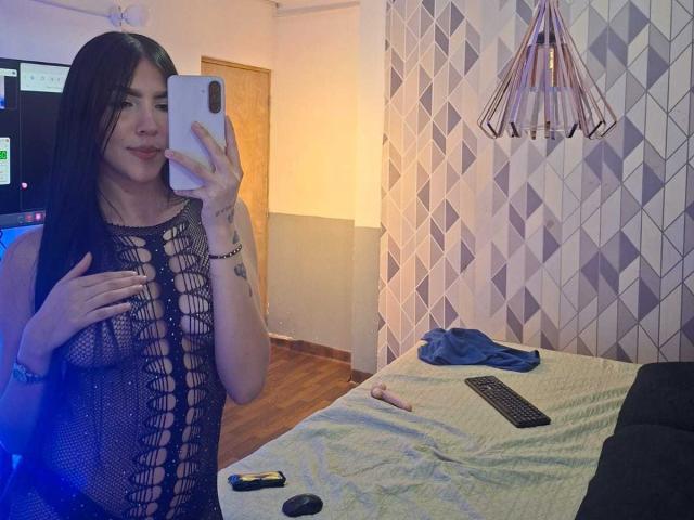La princesse Victoria, avec ses longs cheveux noirs, porte une robe en maille noire qui révèle ses seins. Elle prend un selfie devant un miroir dans une pièce où l'on voit un lit, un téléphone, une télécommande et un sextoy..