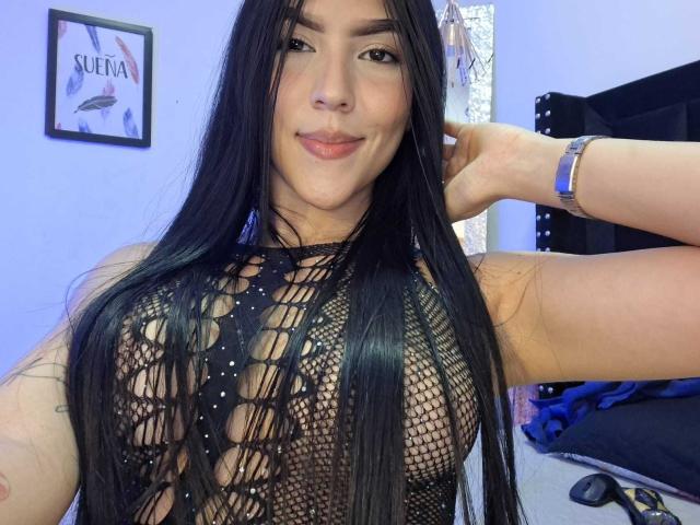 La principessa Victoria, con lunghi capelli neri, indossa una maglia nera a rete che rivela il suo seno prosperoso, sorride alla fotocamera, con il braccio destro alzato, un orologio blu al polso, e alle spalle si vedono pareti viola..