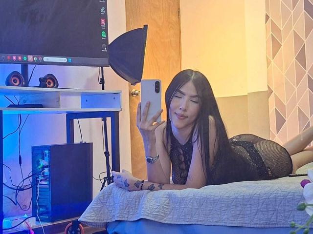 La principessa Victoria, sdraiata su un letto, indossa un abito nero in pizzo, sta scattando un selfie con un telefono, ha lunghi capelli neri, sta sorridendo, ed è accanto a un televisore e a un'attrezzatura per le riprese..