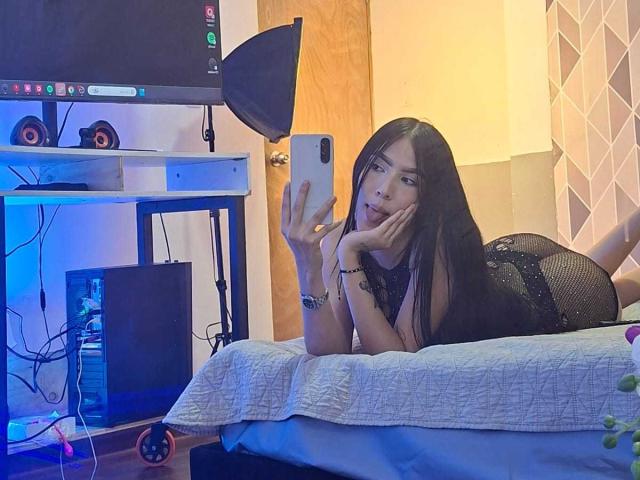 La principessa Victoria, con i suoi lunghi capelli neri, è sdraiata su un letto, indossa un abito di rete nera, tiene in mano un telefono, lo sta guardando, con una mano appoggiata al mento, in una camera da letto..