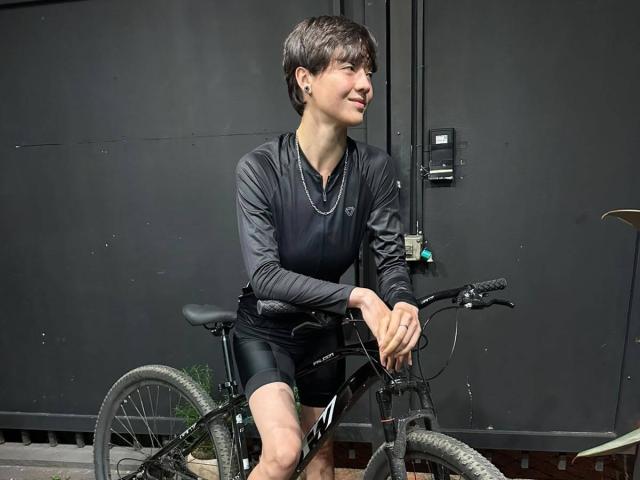 LissyLuxe, com cabelo castanho curto, vestindo uma camisa preta de manga comprida e shorts pretos, andando em uma bicicleta de montanha preta, sorrindo levemente..