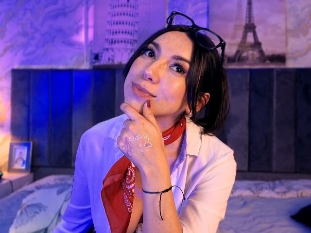 MiaReed with glasses on head, red bandana, white shirt, hand on chin, blue-lit bedroom, black headboard, Eiffel Tower poster, tattoo on hand, slightly pouty expression.