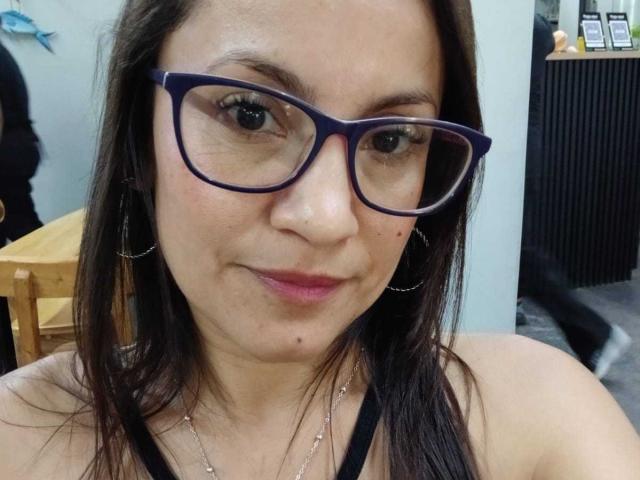 SolaraDubois with dark brown hair, wearing blue glasses and black top, has pink lipstick and small mole under her right eye, smiling slightly, showing a cute face.