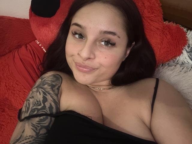 LucieRed with dark red hair, green eyes, and tattoos on her right arm, smiling at the camera, wearing a black top, showing her large tits. Red pillow behind her.