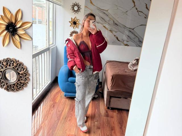 Elena Saenz, met een rode jas, grijze versleten jeans en witte sneakers, die haar kleine borsten laat zien in een spiegelzelfportret, terwijl ze een telefoon vasthoudt, in een kamer met houten vloeren..
