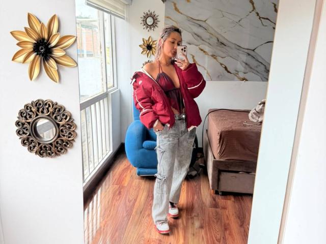 ElenaSaenz with red jacket, grey pants, white sneakers, and red bra, showing her tits, taking a mirror selfie in a room with blue chair and wooden floor.
