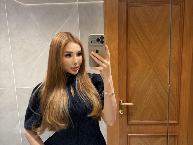 AikoSin, avec de longs cheveux blonds, portant une robe bleu foncé, prend un selfie dans une salle de bain, tenant un iPhone, une porte en bois est visible en arrière-plan, elle regarde l'appareil photo..