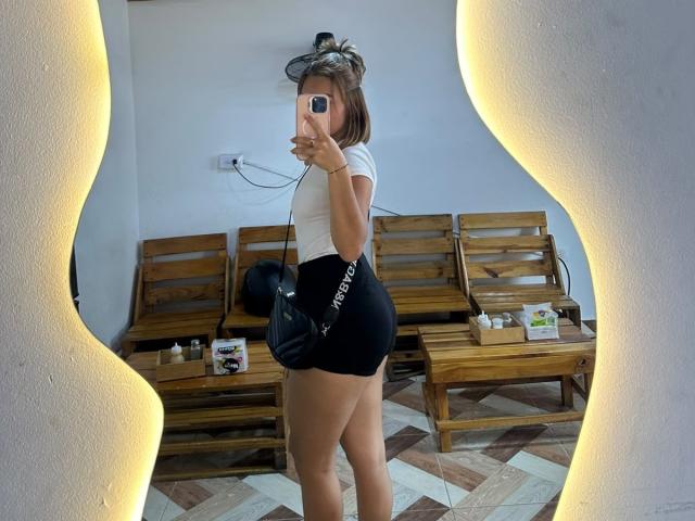 VictoriaSanzF in een witte top, zwarte shorts, met een telefoon in haar hand, die haar achterwerk laat zien, staand voor een spiegel, met een zwarte tas, een houten bankje en een vloer met een patroon op de achtergrond..