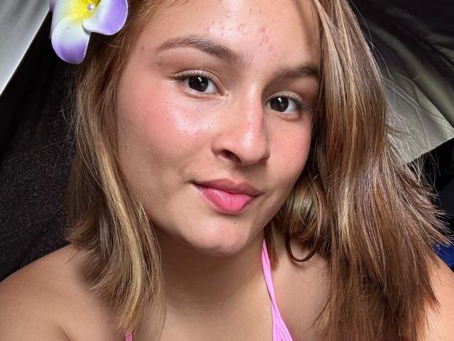 VictoriaSanzF, com cabelo castanho claro, batom rosa e uma flor roxa no cabelo, mostrando um rosto levemente avermelhado com pequenas espinhas na testa, vestindo um top rosa de alças..