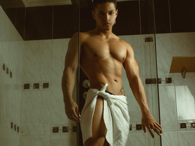 RyanGreyss è in bagno, senza camicia, con un asciugamano bianco avvolto intorno ai genitali, mostrando un sedere muscoloso e tonico, e un'espressione seria. Ha i capelli scuri e la pelle marrone..