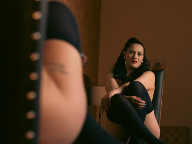 Elizabeth Dupont, con i capelli neri, il rossetto rosso e le calze nere che arrivano all'altezza coscia, seduta su una sedia, con le gambe incrociate, mostra i suoi seni nudi, guardando uno specchio che riflette il suo sedere..