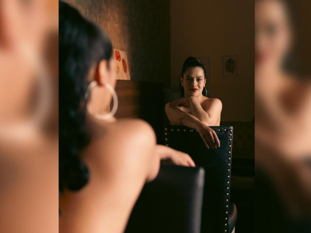 Elizabeth Dupont, con i capelli neri raccolti in una coda di cavallo, indossa grandi orecchini a cerchio e siede nuda di fronte a uno specchio, con le braccia incrociate, osservando il suo riflesso, in una stanza scarsamente illuminata..