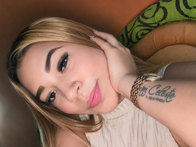 Aisha, con cabello rubio, cejas marrones, lápiz labial rosa, piercing en la nariz, blusa blanca, mano en la mejilla, tatuaje en la muñeca, pulsera y uñas rosas, mirando a la cámara..