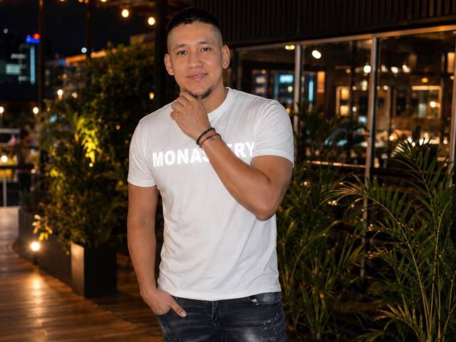 DanteVega mit kurzen, schwarzen Haaren, gebräunter Haut und muskulöser Statur, trägt ein weißes T-Shirt mit der Aufschrift "MONDAY", eine schwarze Jeans und ein Armband und steht mit einer Hand unter dem Kinn..