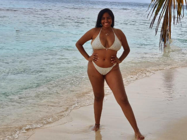 SiennaTaylor stands on a beach in a white bikini, hands on hips, smiling with dark skin and curly black hair, showing her big tits and curvy ass.