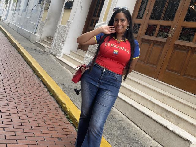 SiennaTaylor with brown skin, long black hair, red and blue shirt, blue jeans, holding phone, smiling, standing on brick sidewalk, yellow curb, wooden doors, red purse, earrings.