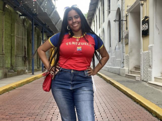 SiennaTaylor has big boobs, long black hair, and is smiling. She's wearing a red and blue Venizelles shirt, tight jeans, and a red bag, standing on a brick sidewalk.