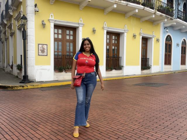 SiennaTaylor has dark skin, long black hair, red t-shirt, blue jeans, yellow shoes, red crossbody bag, standing on brick sidewalk, yellow and white building in background.