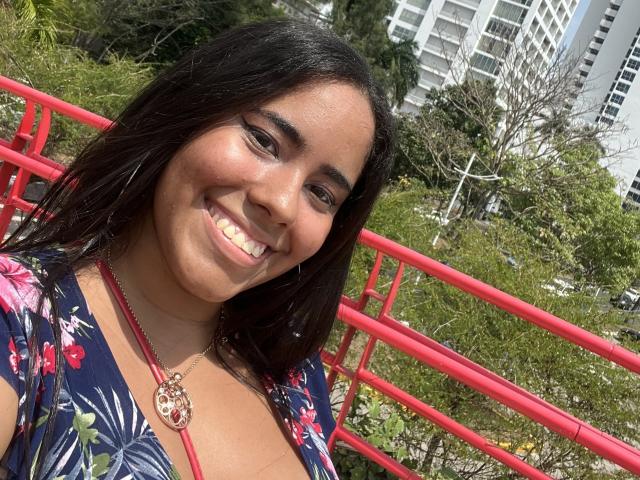 SiennaTaylor with dark brown hair, smiling, wearing a floral blue dress, red necklace, and red earrings, standing in front of a red railing, sunny day, green background.