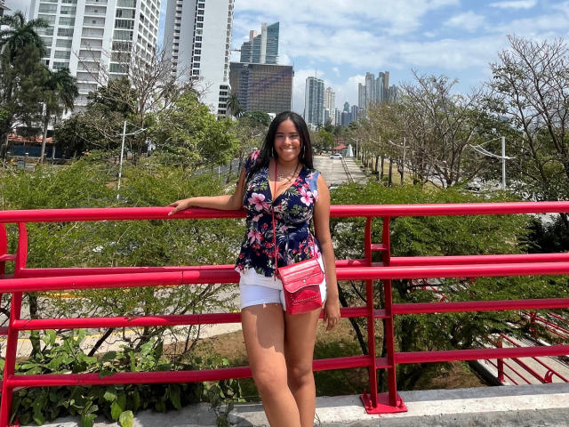 SiennaTaylor with medium tits, wearing a floral shirt, white shorts, and pink crossbody bag, leans on a red railing, smiling, with green trees and tall buildings in the background.