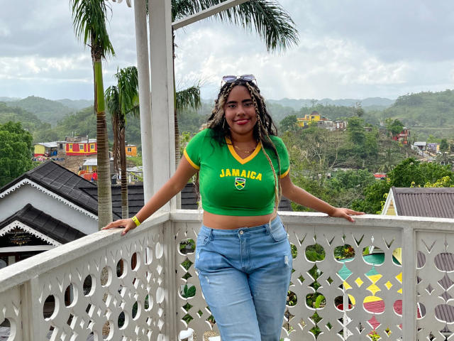 SiennaTaylor with big tits, green crop top, yellow trim, blue jeans, standing on white balcony, palm tree, cloudy sky, mountains, and houses in background, smiling, black braided hair.