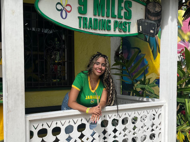SiennaTaylor with long braided hair, green shirt, yellow trim, leaning on white lattice, smiling, wearing blue jeans, sign above reading "9 Miles Trading Post".