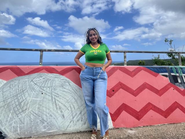 SiennaTaylor with curly blue-gray hair, green crop top, and blue jeans, leaning against a red and white wall, hands in pockets, smiling, under a cloudy blue sky.